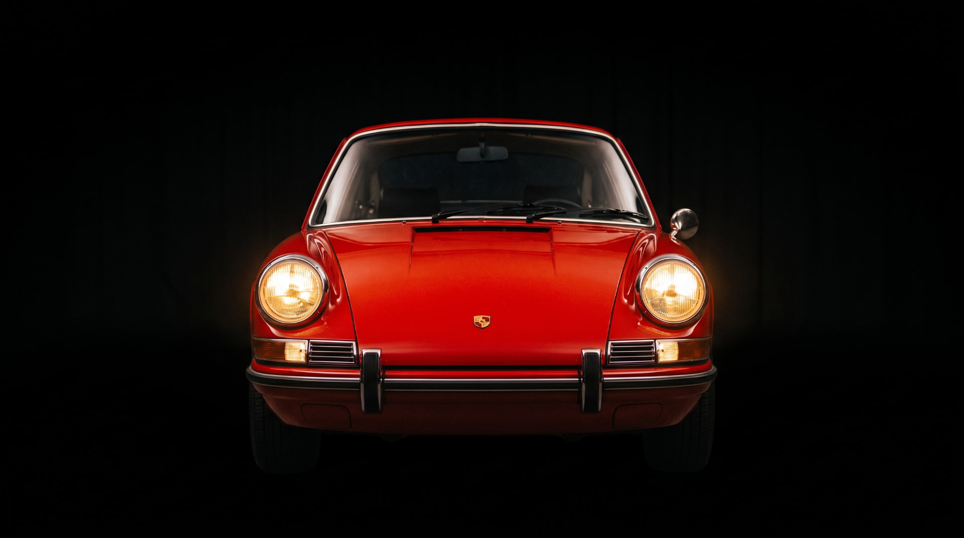 Porsche 911 d'epoca rossa vista di fronte, con i fari accesi su sfondo nero